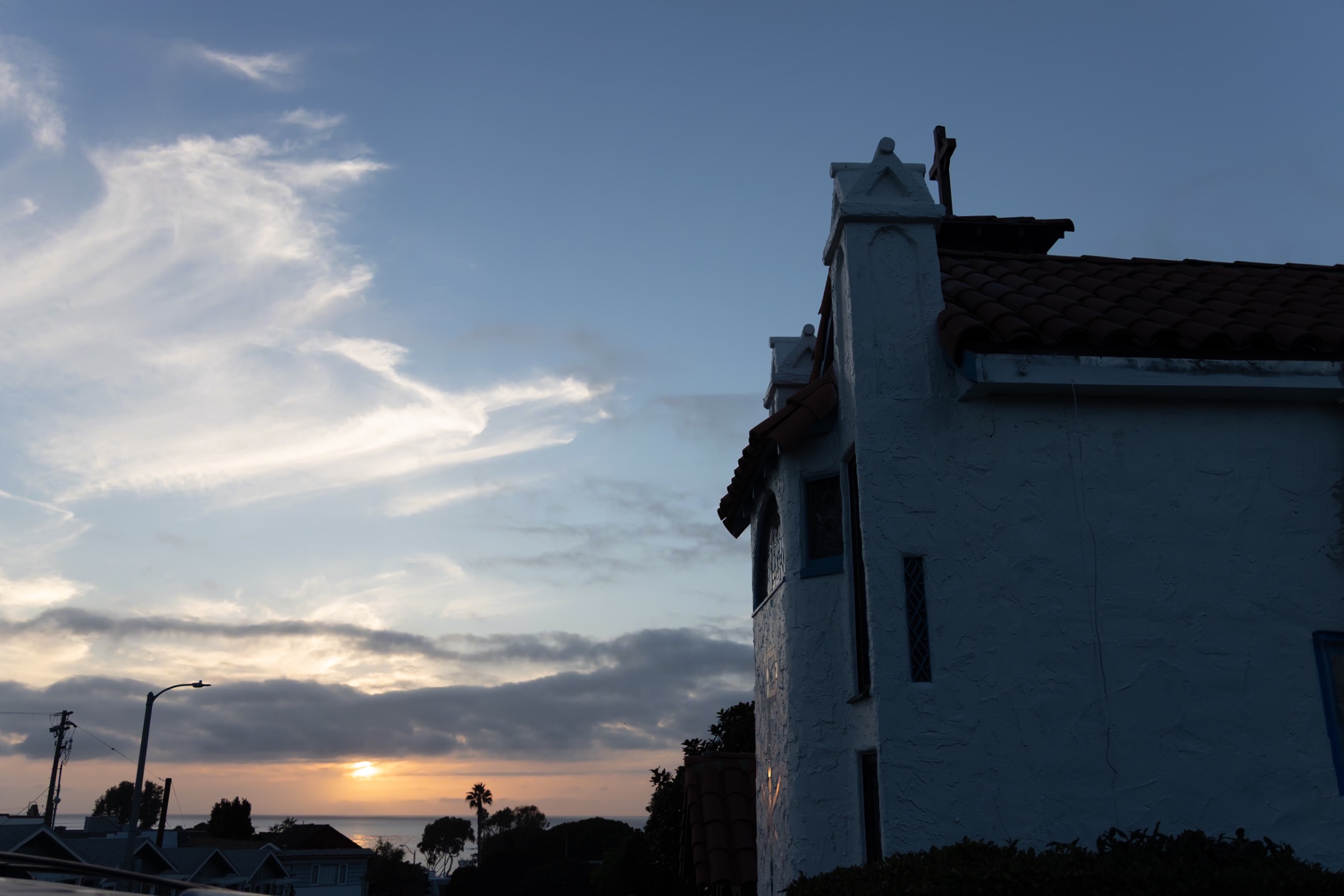 Side of St. Francis by-the-Sea at sunset: white stucco walls, a red clay tile roof and a cross on a gabled pier, with the sun setting over the Pacific beyond rooftops and palm trees.