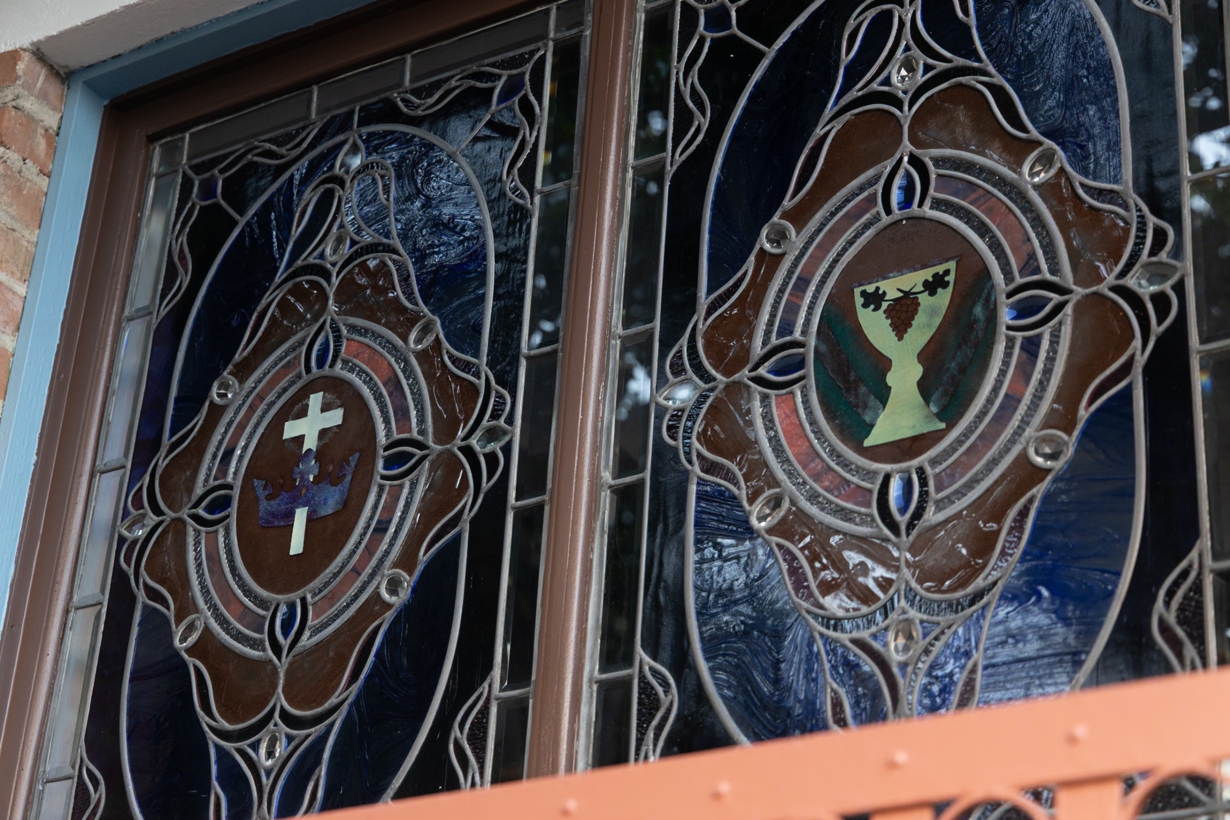 Close view of two leaded stained-glass panels in deep blue: one shows a white cross rising through a crown, the other a gold chalice with a cluster of grapes and vine leaves.