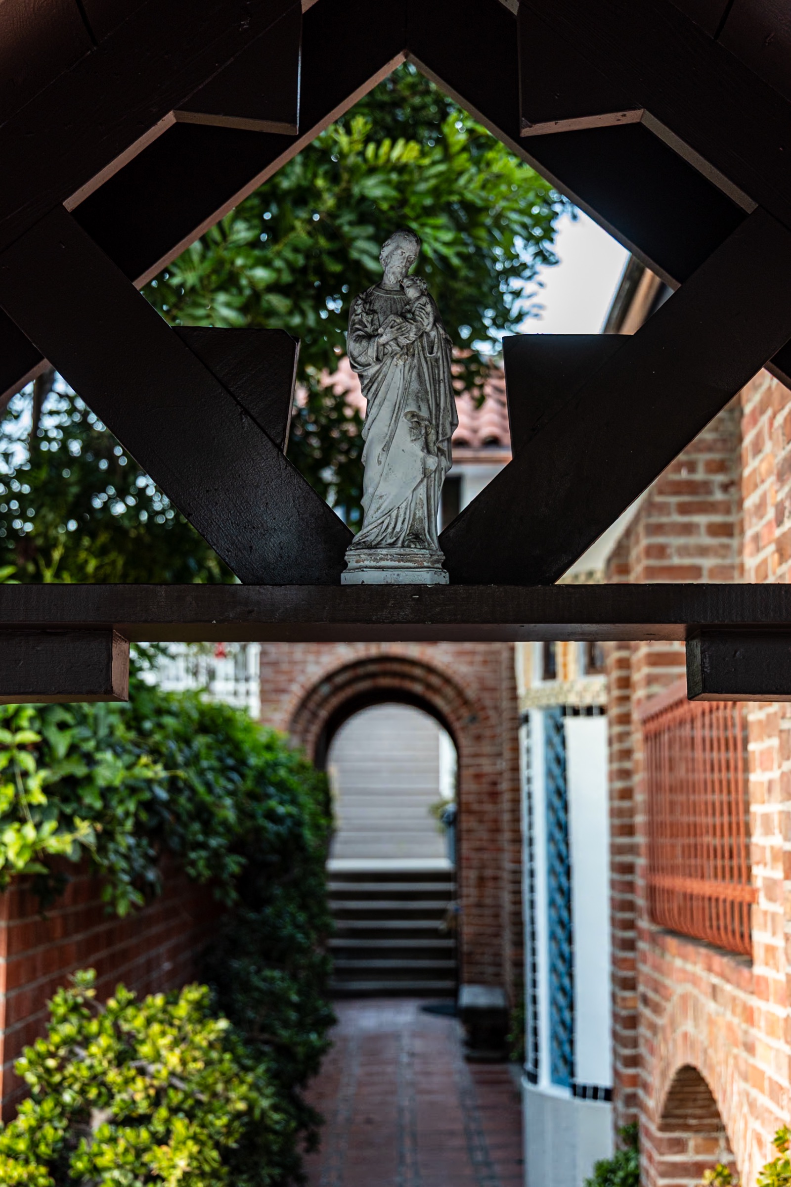 Weathered white statue of St. Joseph holding the Christ Child, standing on a dark timber beam above a brick walkway that leads to an arched brick doorway and steps.